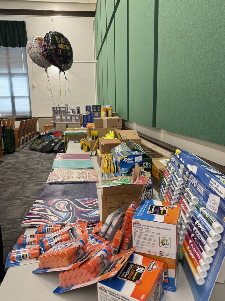 A close-up view of tables filled with donated school supplies, including notebooks, glue sticks, markers, crayons, pencils, folders, and other classroom materials.