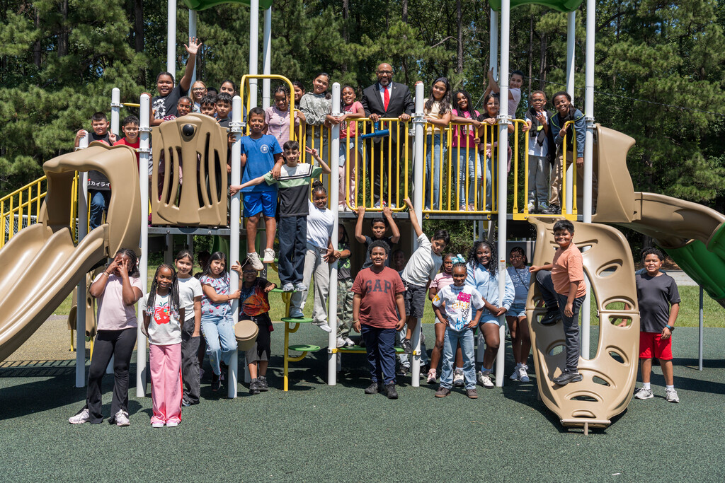 A diverse group of children and adults smile on a tan-and-yellow playground with slides and climbing bars, surrounded by trees in bright sunlight.