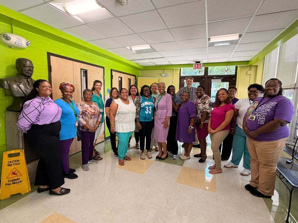 Group photo of school staff posing together in a school hallway for Teal & Purple Spirit Day. Several staff members wear teal and purple clothing while smiling and celebrating school spirit, connection, and unity.