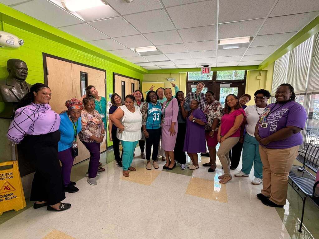 Group photo of school staff gathered in a hallway wearing teal, purple, and other colorful clothing for Teal & Purple Spirit Day. Staff members smile and pose together in a show of school spirit and unity.