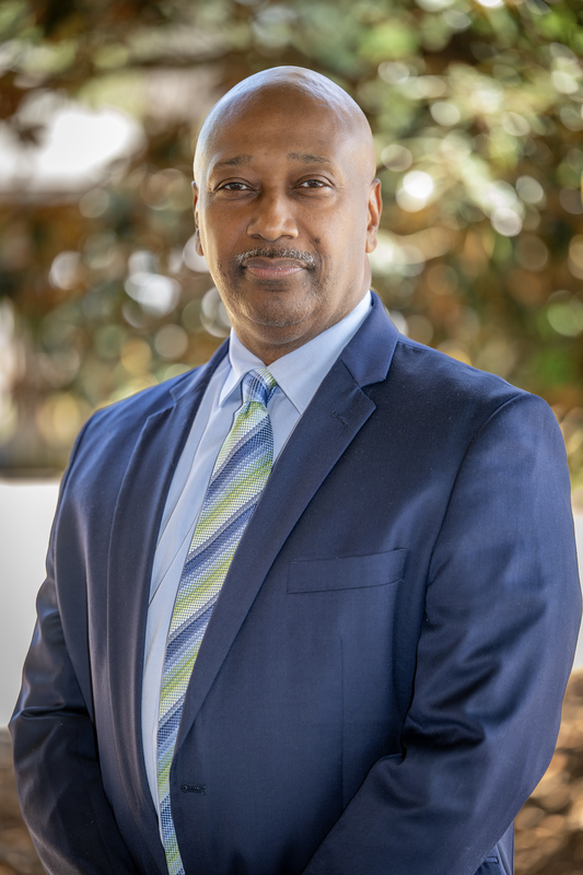 Dr. Hooker in a blue suit and patterned tie stands outdoors, smiling confidently. The background is blurred greenery, creating a professional yet relaxed tone.