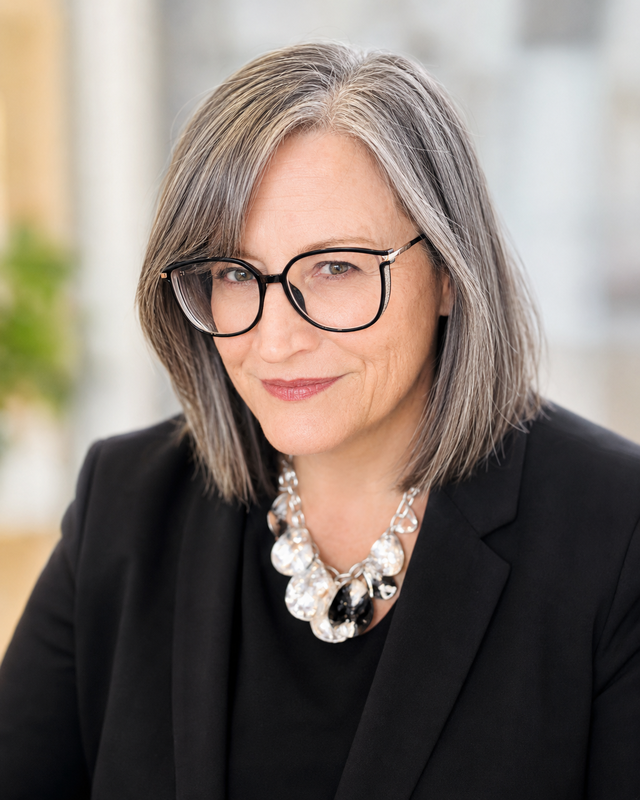A woman with glasses and gray hair, wearing a black suit, smiles slightly.