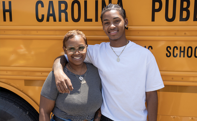 A young man and woman pose happily in front of a school bus, capturing a fun moment together.