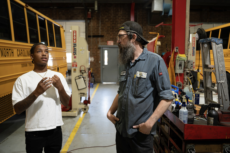 Two men are in a garage with yellow school buses in the background. One in a white shirt is talking, while the other in a uniform listens intently.