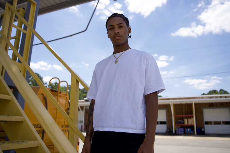 A young man stands confidently outdoors under a blue sky, wearing a white t-shirt and gold necklace. Yellow stairs and an industrial building are in the background.