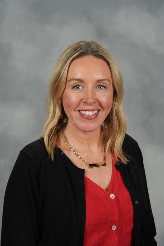 Smiling woman with blonde hair wearing a red blouse and black cardigan. She has a gold necklace. Gray background, professional and approachable tone.