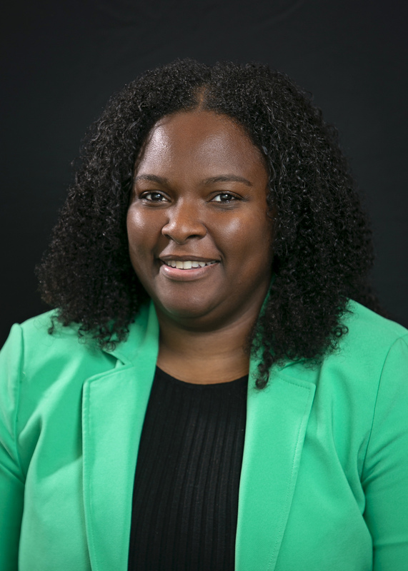 A person with curly hair smiles at the camera against a dark background. They are wearing a bright green blazer over a black top, conveying a professional yet approachable look.