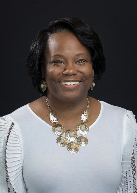Smiling woman wearing a white blouse and gold jewelry, standing against a black background. Her expression is warm and welcoming.