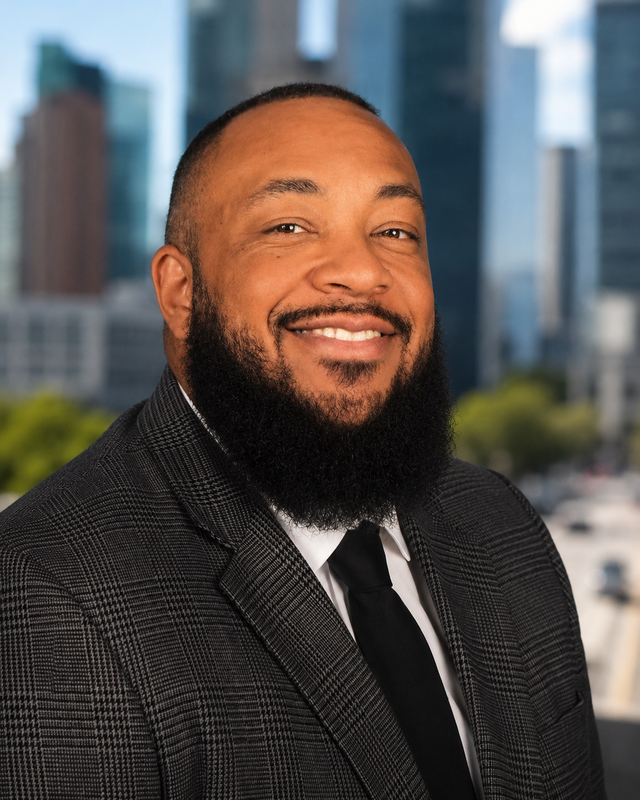 Professional headshot of a smiling man with a full beard, wearing a gray plaid suit and black tie against a blurred city background.