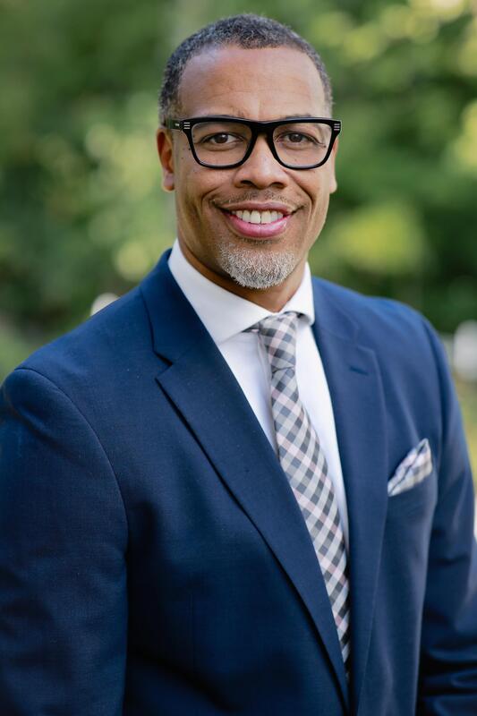 Professional headshot of a smiling man wearing glasses and a navy suit with a patterned tie, standing outdoors against a blurred green background.