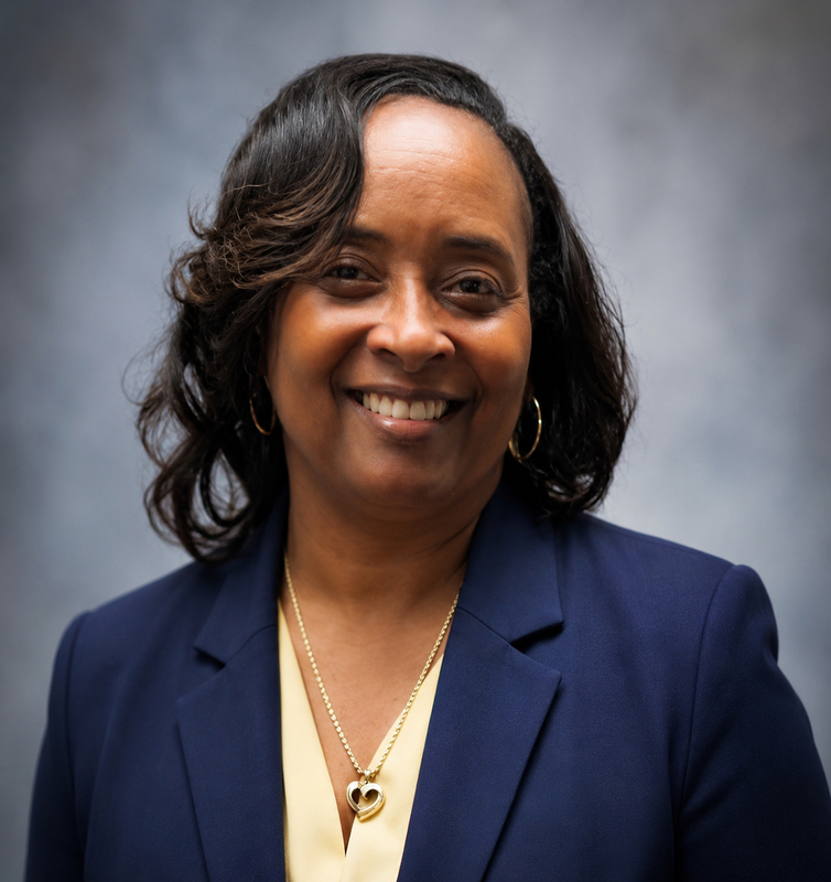 Professional headshot of a smiling woman wearing a navy blazer and gold necklace against a soft gray background.