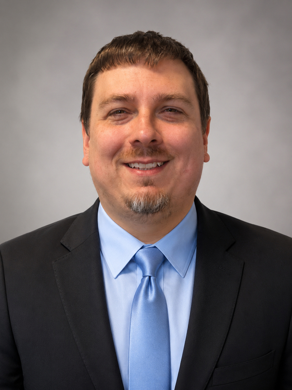 Professional headshot of a smiling man wearing a black suit, light blue shirt, and blue tie against a gray background.