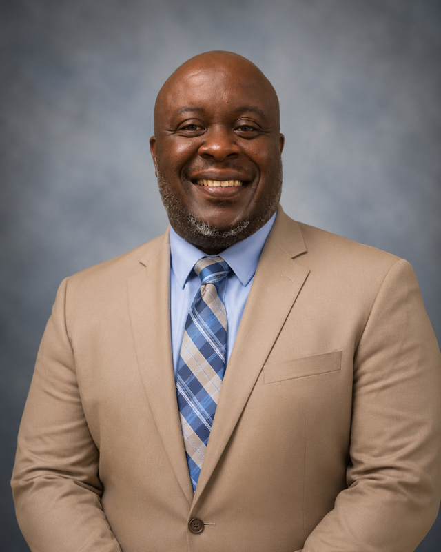 Professional headshot of a smiling man wearing a tan suit and blue plaid tie against a blue-gray background.