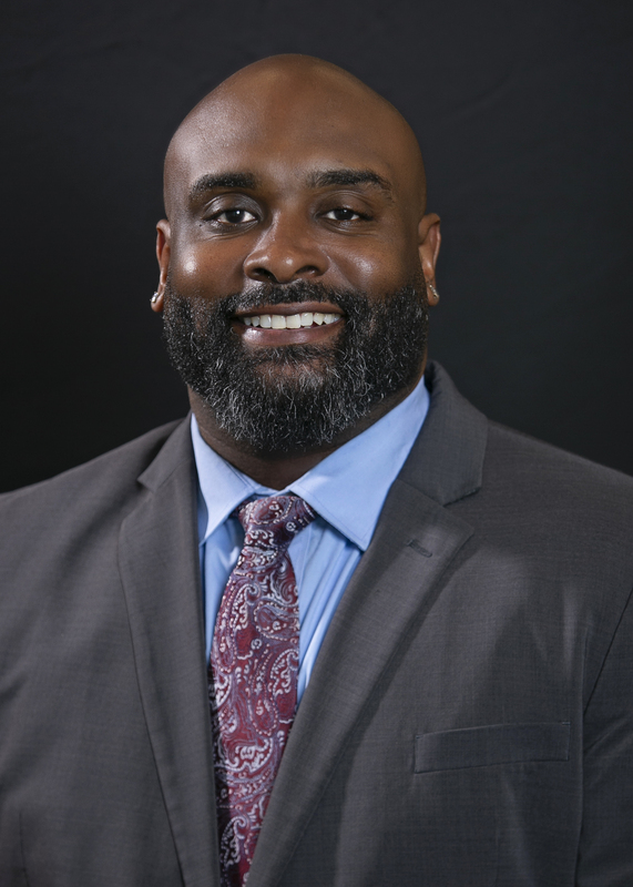 Professional headshot of a smiling man with a beard, wearing a charcoal suit, blue shirt, and patterned tie against a black background.