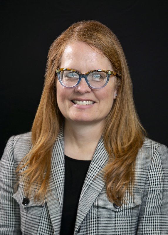 Professional headshot of a smiling woman with red hair and glasses, wearing a patterned blazer against a black background.