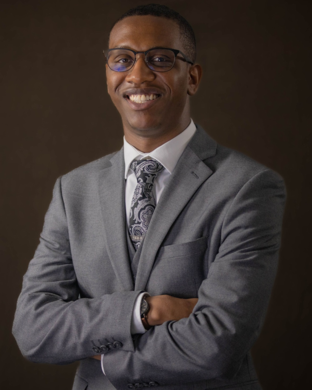 Smiling man in glasses and a gray suit stands with arms crossed against a dark background, conveying confidence and warmth in a professional portrait.