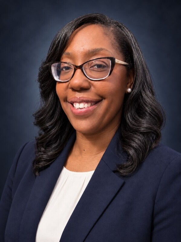 A smiling Black woman in glasses, a white blouse, and navy blazer poses against a dark studio backdrop, conveying warmth and confidence.