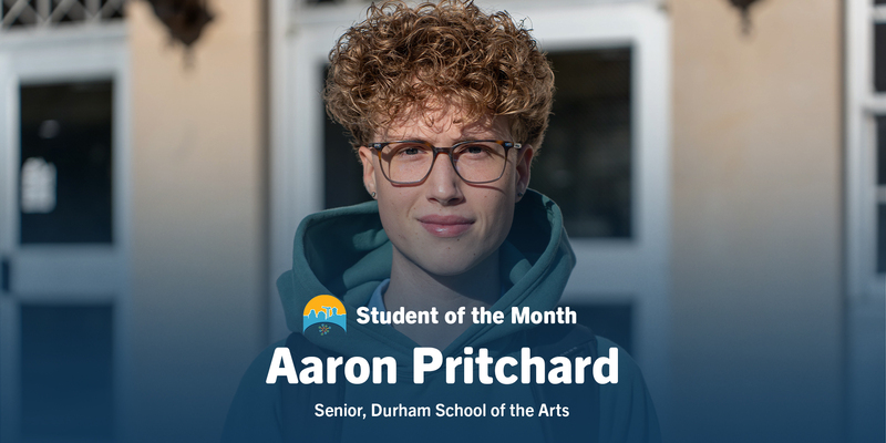 Student of the Month Aaron Pritchard, a curly-haired senior in glasses and a teal hoodie, smiles outside Durham School of the Arts.