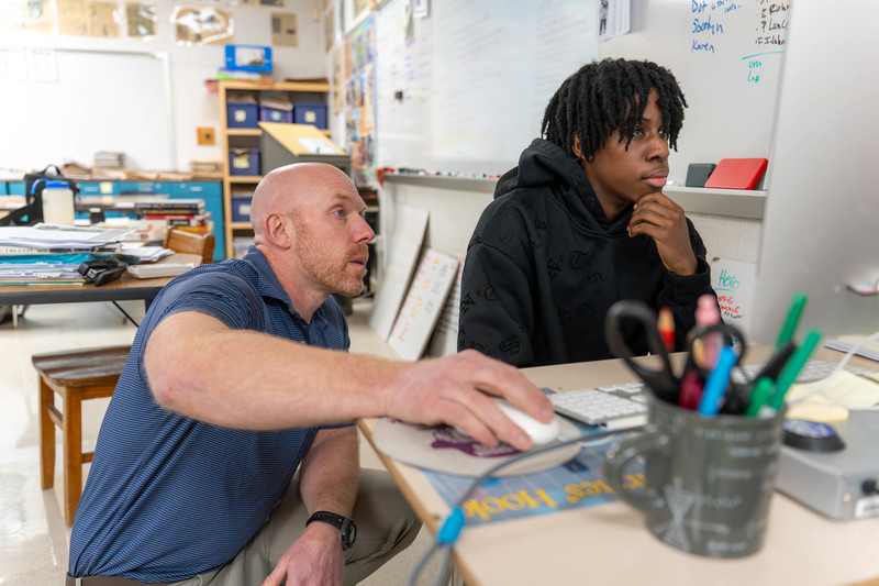 A teacher and student engage at a computer in a classroom. The teacher points at the screen while kneeling, the student focuses, hand on chin. Papers, supplies, and notes surround them, suggesting a collaborative learning environment.