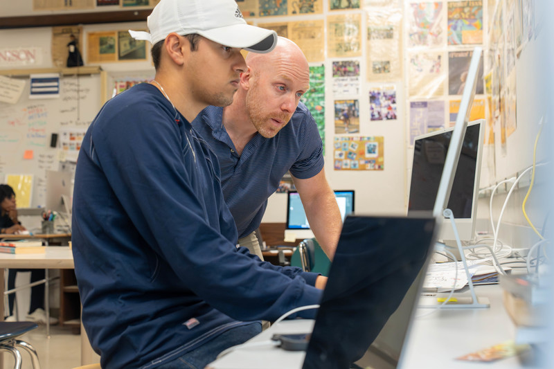 Two men in a classroom setting work at a computer. One in a cap is seated, focused on the screen, while the other leans in, attentively assisting.