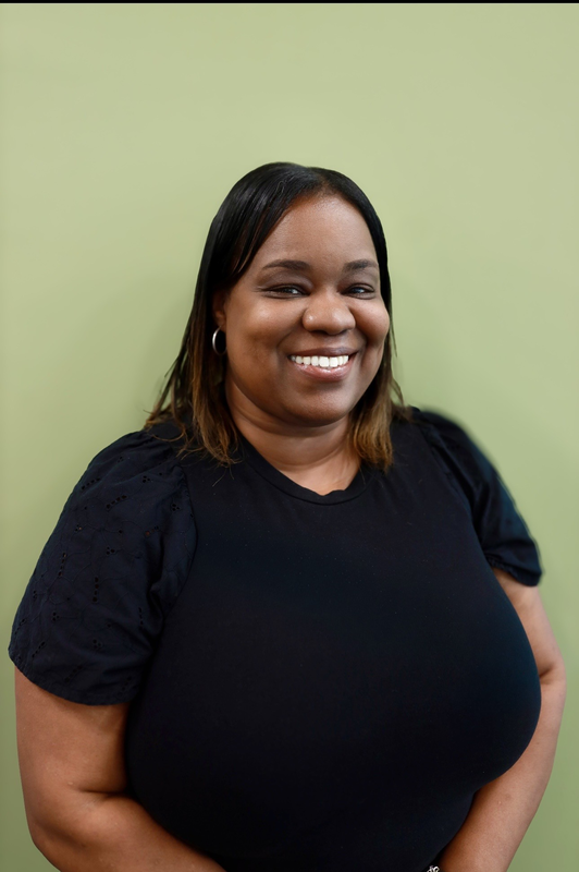 Smiling woman in a black shirt stands against a light green background. Her hair is shoulder-length, and she appears content and approachable.