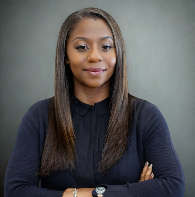 Confident woman with straight hair smiling, wearing a dark top with arms crossed against a neutral gray background, conveys professionalism.