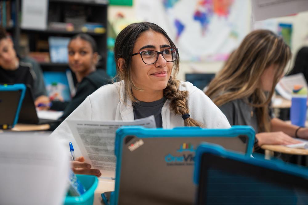 A smiling female student is seated at her desk in the classroom  and is holding classwork in her hand. 