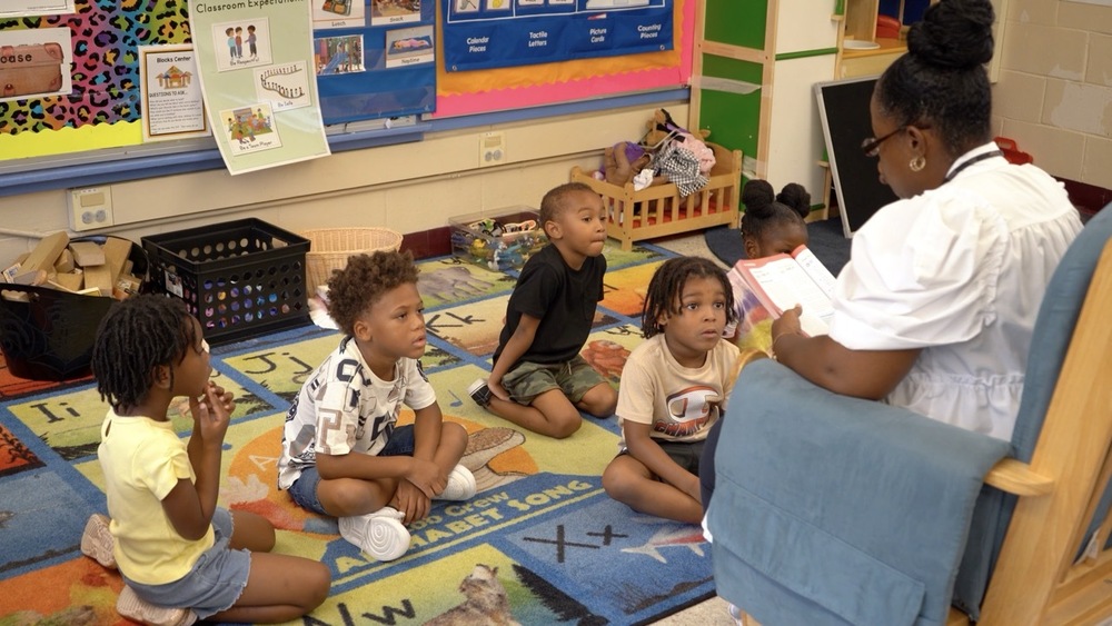 A group of students seated on a carpet listen to a VPK teacher reading a story.