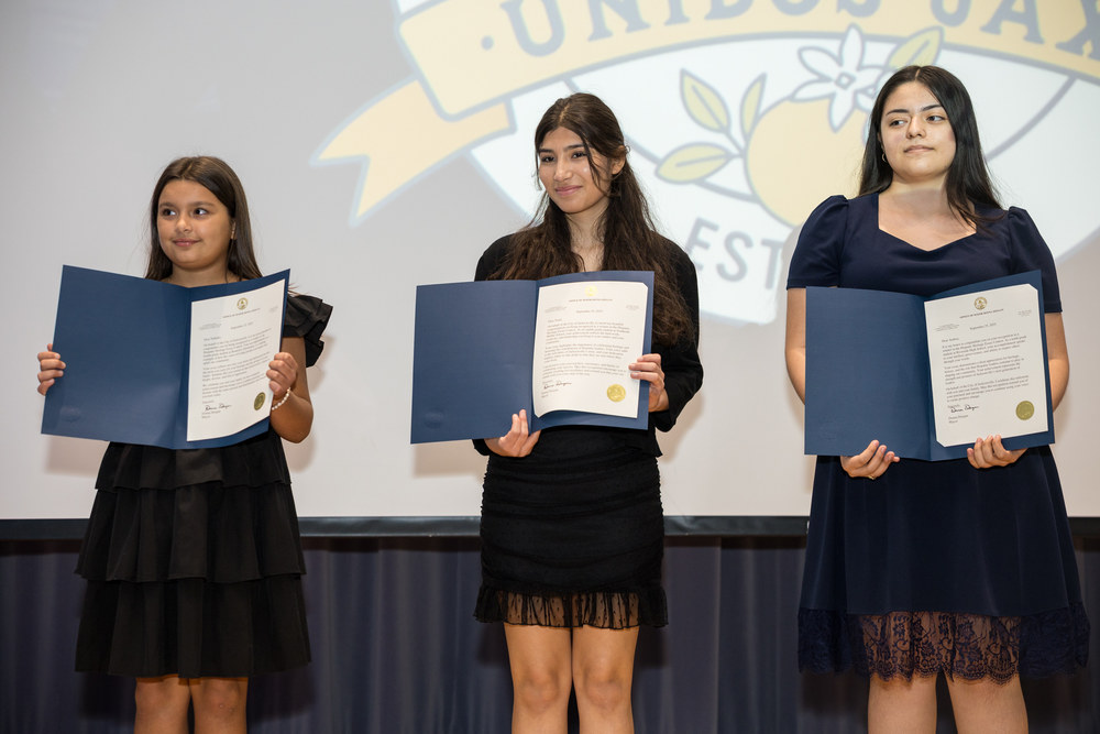 The three winners of the essay contest stand on stage during a ceremony and display certificates.