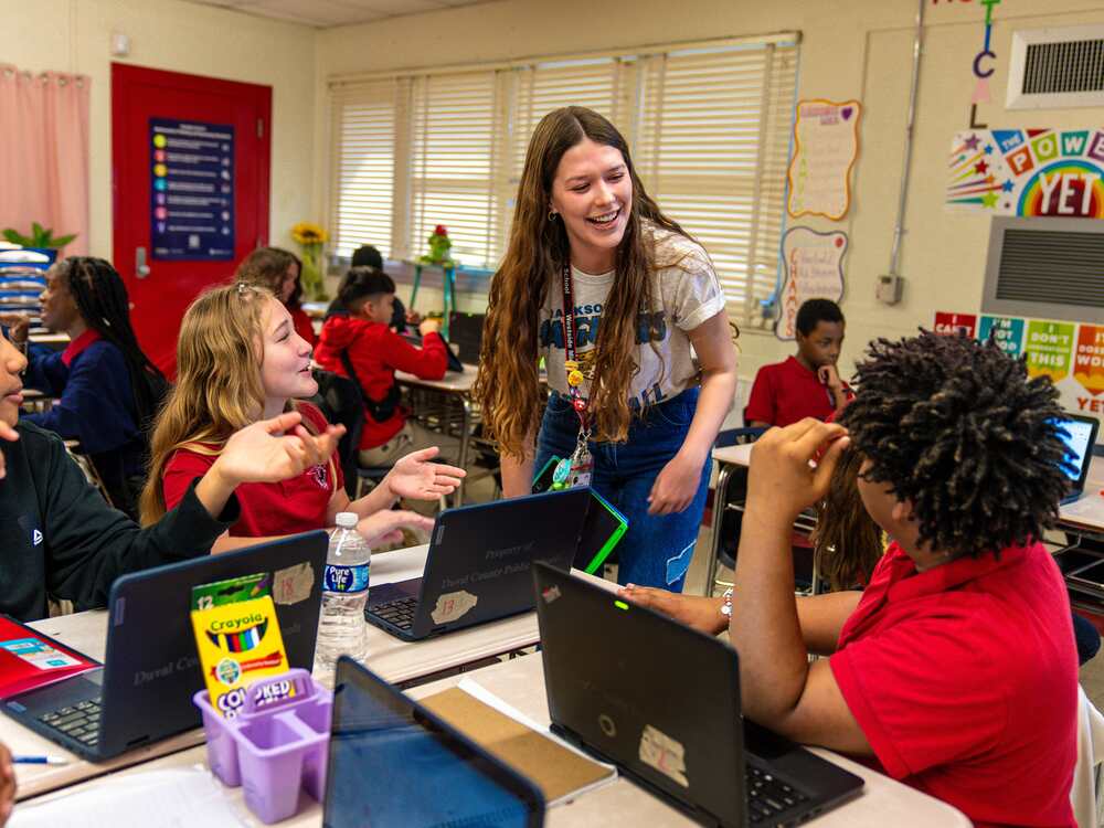 A teacher smiles at two students in the classroom as they work on the laptops