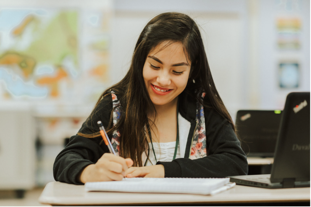An image of a  girl happily writing study notes at her desk