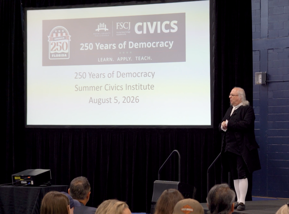 A Ben Franklin impersonator stands at the front of an auditorium, addressing a crowd of social studies teachers.