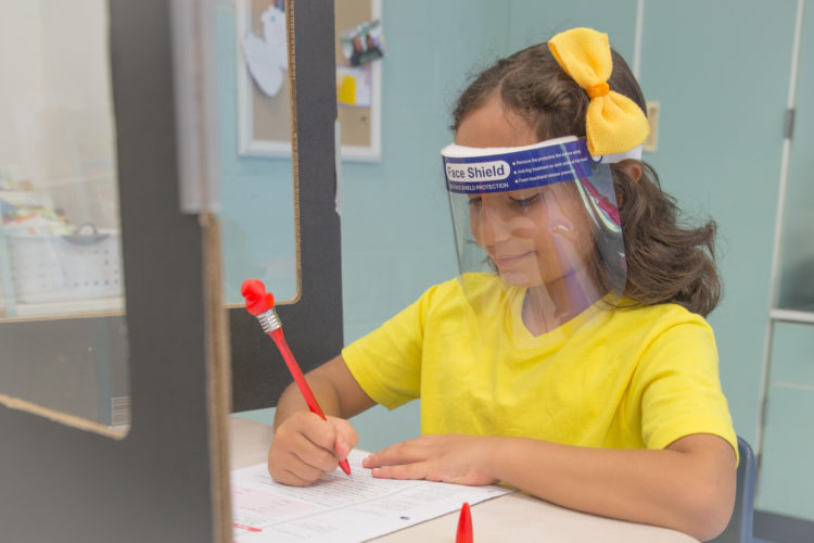 Student writing at desk, surrounded by a  desk divider and wearing a face shield for Covid-19 precautions 