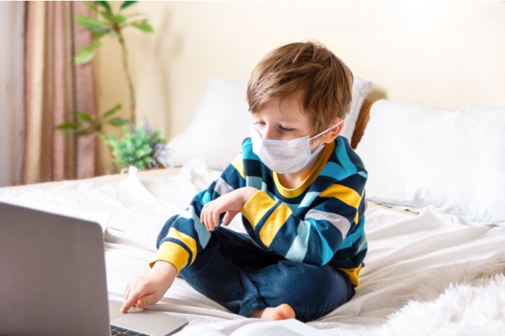 Image of a little boy in a mask, sitting on the bed working on a laptop