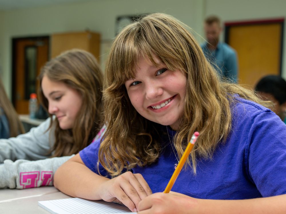 A student sits at a desk with a pencil in her hand and smiles at the camera.