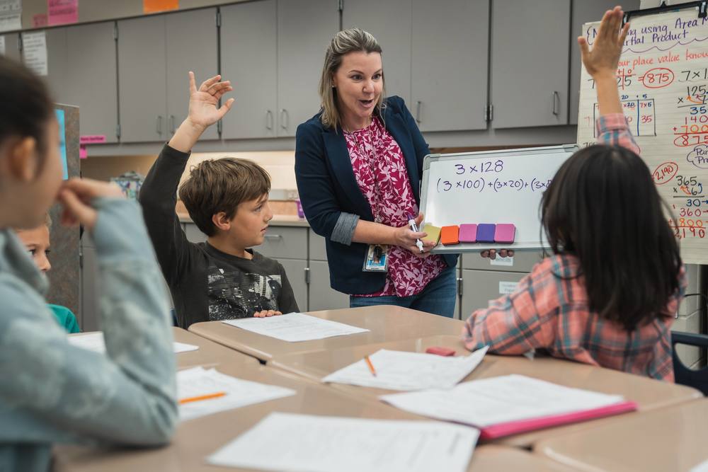 Students raise their hands as their teacher shows off a white board