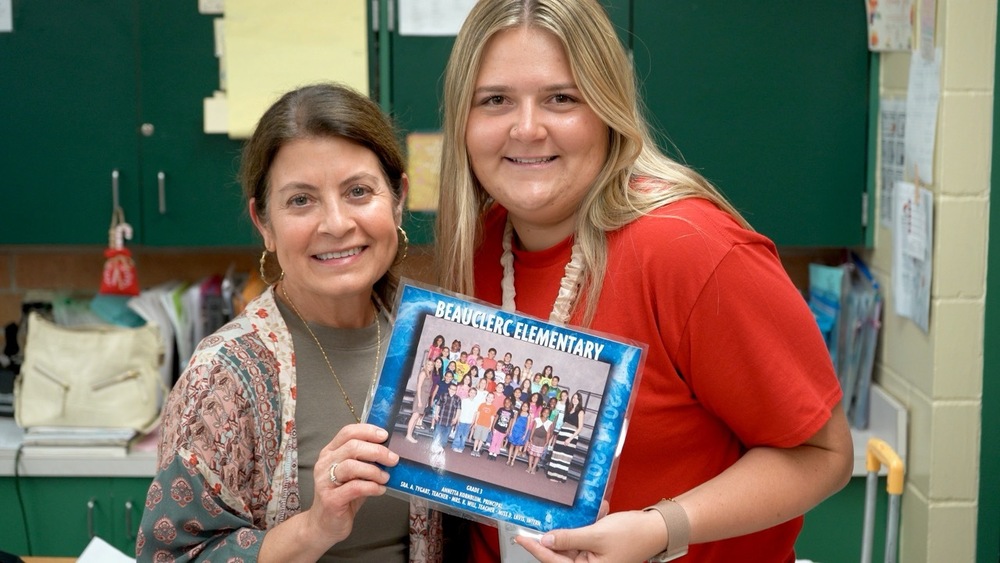 Two teachers stand together in a classroom, holding an old class photo.