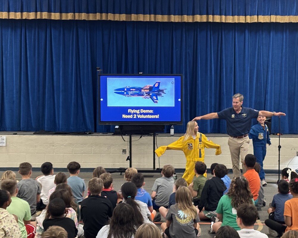 A man and a boy and a girl standing in front of a stage and a digital monitor with a blue angel fighter jet flying in the air. The girl is in a yellow flight suit and the boy is in a blue flight suit. The three of them are modeling a flight formation. There are students in front of them, sitting on the floor.
