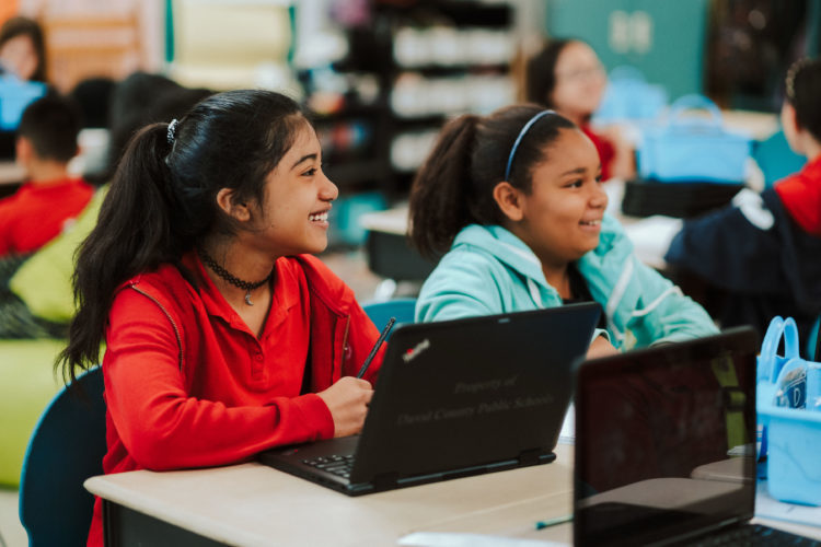 Students smiling in a class room with laptops in front of them. 
