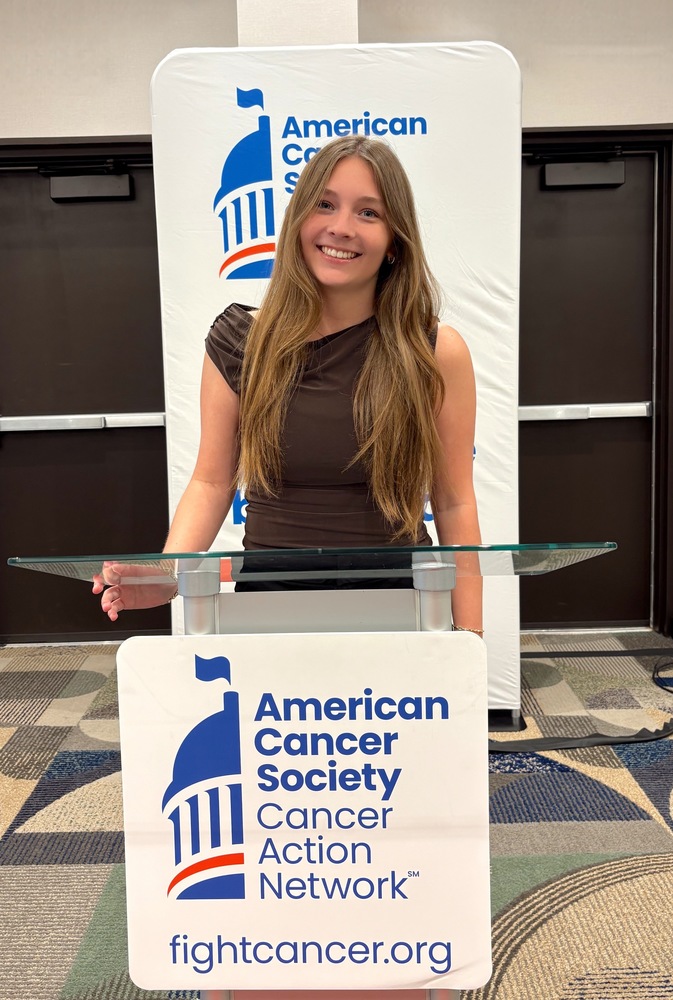 Lidia Ray standing at a lectern in front of an American Cancer Society banner