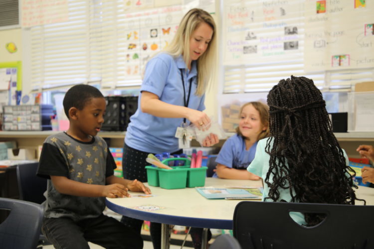 Students sit around a table with craft supplies, while teacher stands, giving supplies out. 