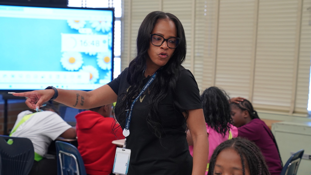 Tahisha Merrell, a 4th grade teacher and U.S. Air Force veteran at Normandy Village Elementary, stands in her classroom smiling as students work at their desks. She's wearing a lanyard and holding student papers, surrounded by colorful educational posters and engaged learners.