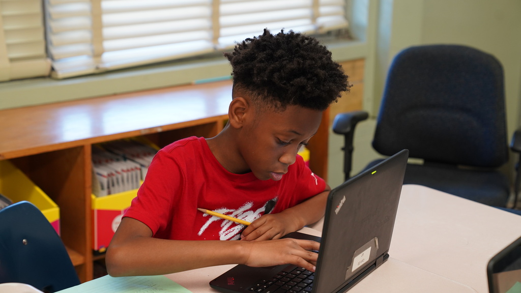 Tahisha Merrell, a 4th grade teacher and U.S. Air Force veteran at Normandy Village Elementary, stands in her classroom smiling as students work at their desks. She's wearing a lanyard and holding student papers, surrounded by colorful educational posters and engaged learners.