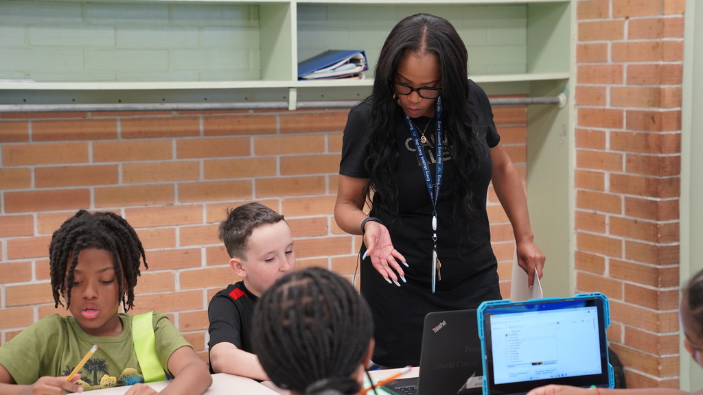 Tahisha Merrell, a 4th grade teacher and U.S. Air Force veteran at Normandy Village Elementary, stands in her classroom smiling as students work at their desks. She's wearing a lanyard and holding student papers, surrounded by colorful educational posters and engaged learners.