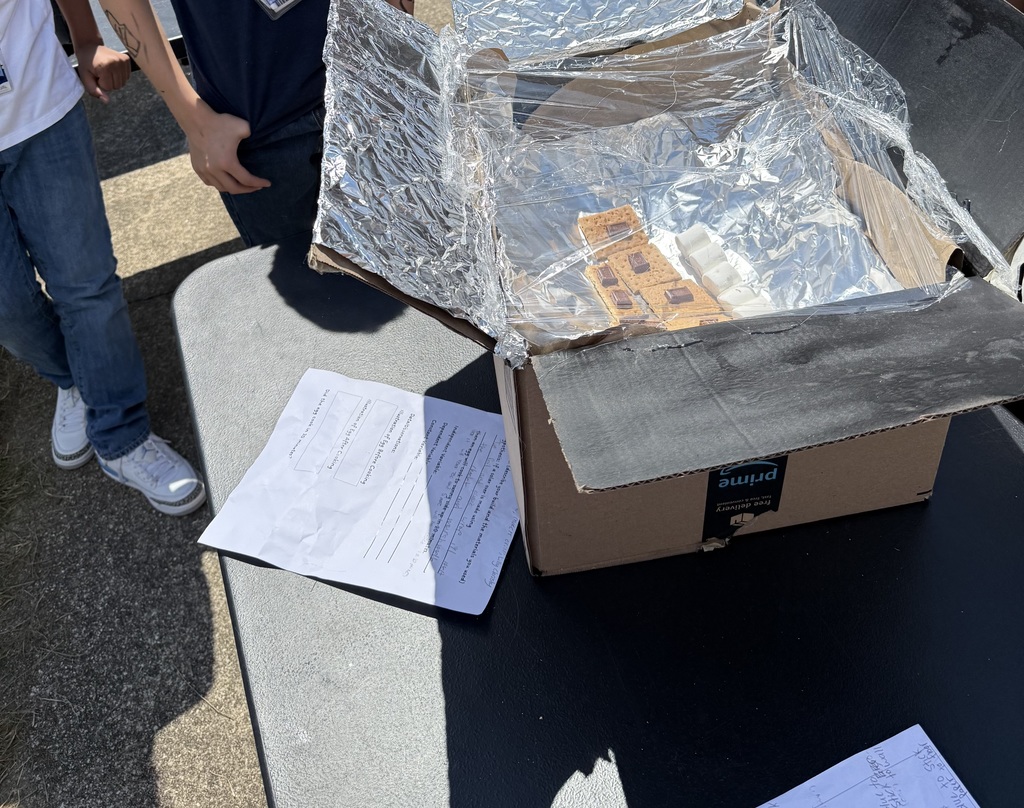 Students at outdoor tables display homemade solar ovens built from cardboard boxes lined with foil, each filled with marshmallows, graham crackers, and chocolate to make s’mores.