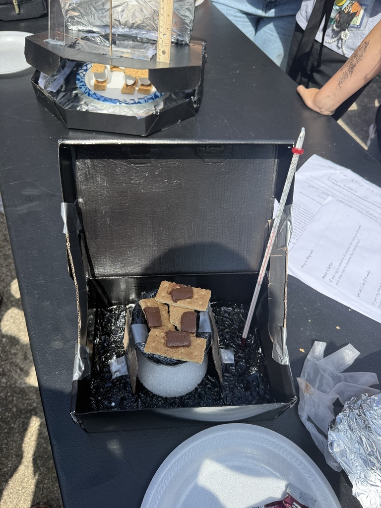 A cardboard box oven covered with plastic wrap holds s’mores ingredients on a paper plate while students stand nearby.
