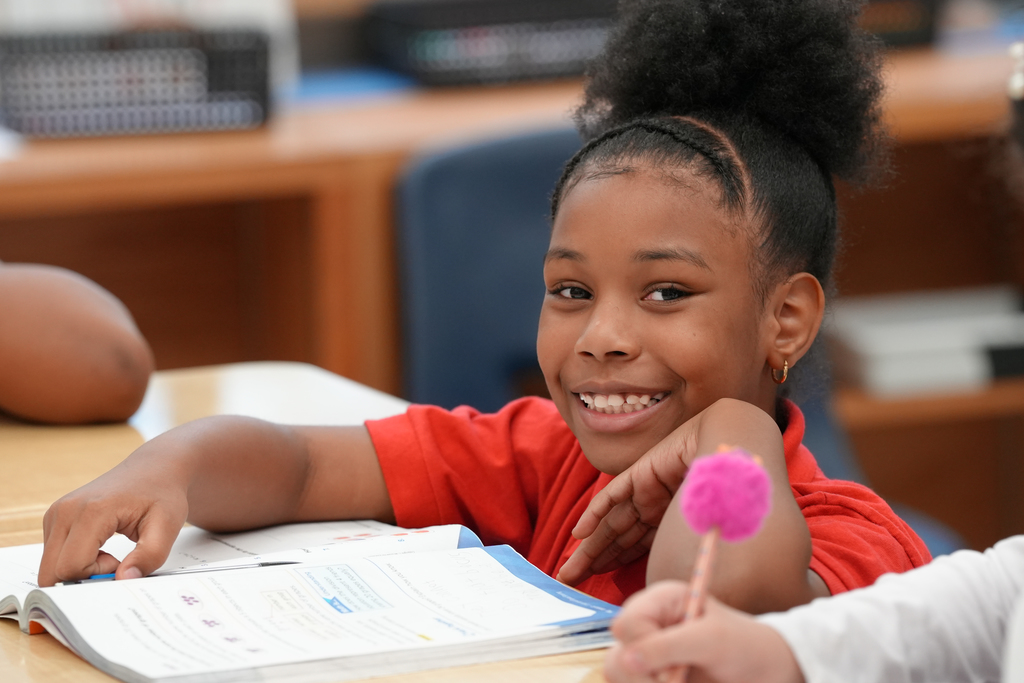 A young girl focuses on her work while a teacher in an orange shirt assists nearby.