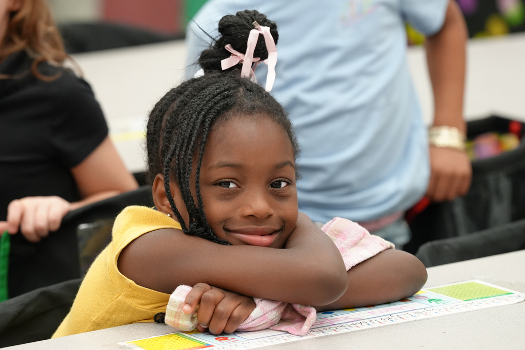 A young girl with braids and a pink bow smiles warmly as she rests her arms on her desk in class.