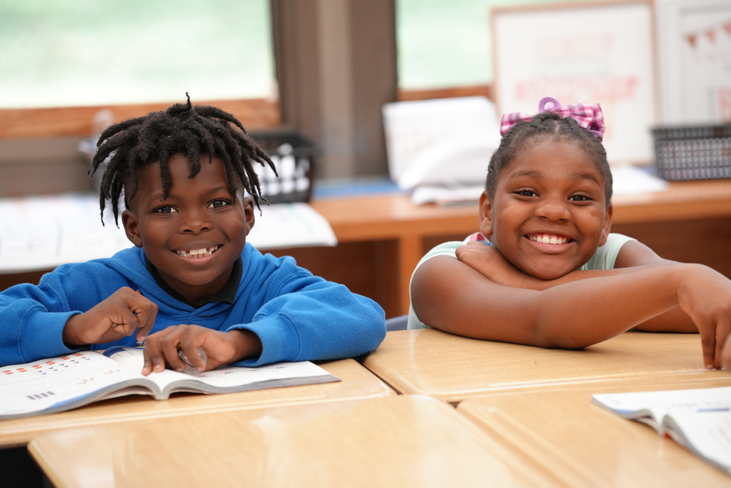 Two smiling students sit close together at a classroom table, leaning toward the camera.