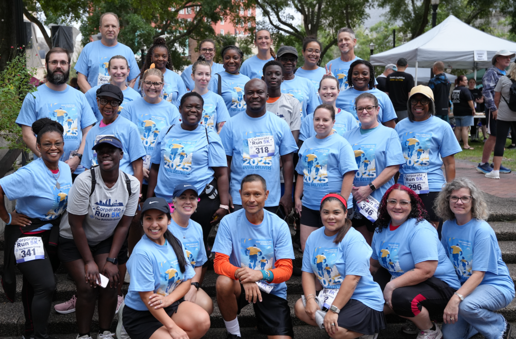 A large group of Duval County Public Schools employees wearing matching blue “Duval Delivers 2025” shirts smile together on a set of outdoor steps before the BKS Corporate Run. The group includes runners and walkers of diverse ages and backgrounds, all showing Team Duval spirit.